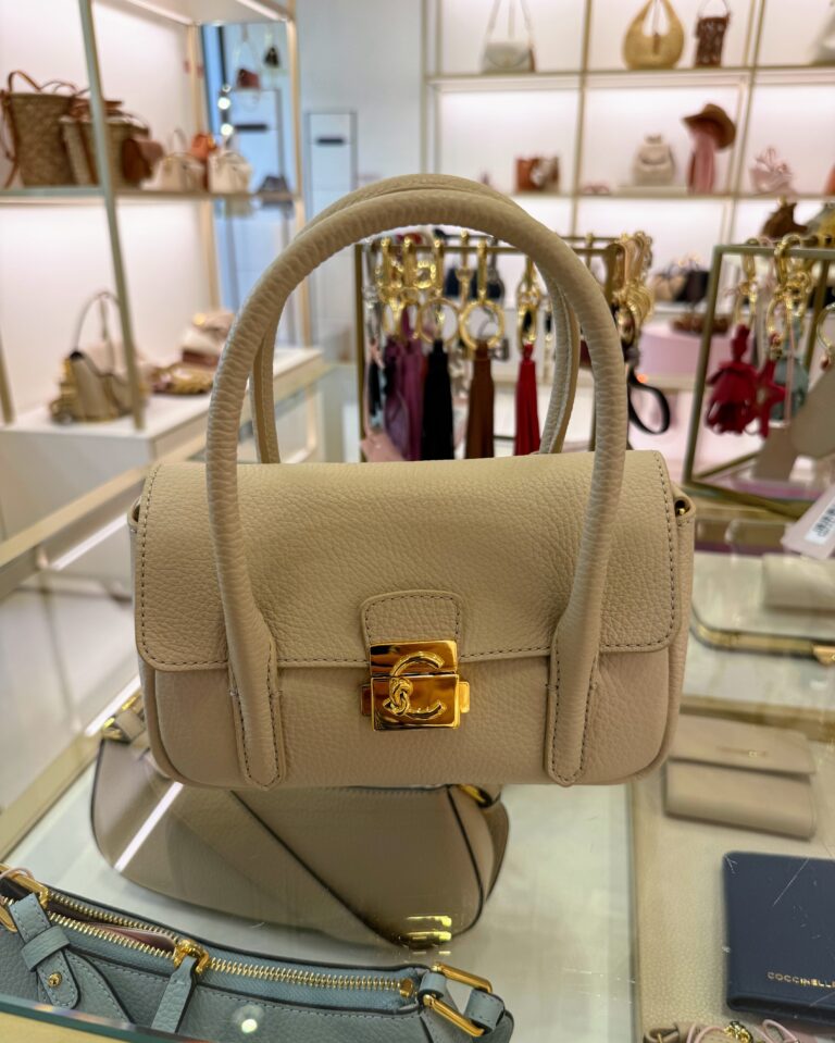 Cream leather handbag with gold clasp on the front that has a big letter C on it for Coccinelle, an Italian leather purse company. The bag is sitting on a glass case with other purses, wallets and keychains.