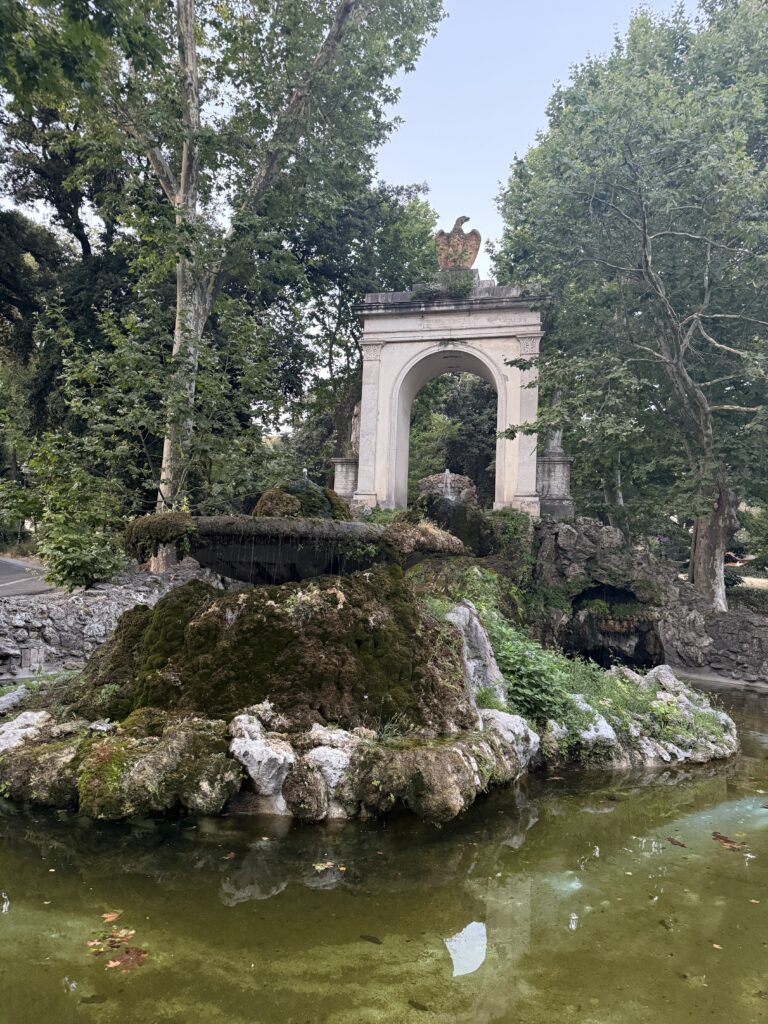 A fountain with mossy rocks and a marble arch behind it with an Eagle statue sitting on the arch. Trees in the background.