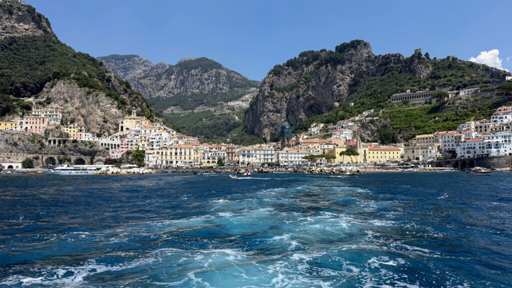 View of Amalfi town from the sea. Colorful buildings along a mountain on a blue sea with boats near the dock.