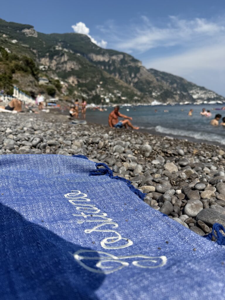 View of Fornillo Beach in Positano Italy. The angle is from below on the corner of a blue towel laid out on the rocks with "Positano" embroidered in white, looking out onto the sea with mountains on the side and ahead.