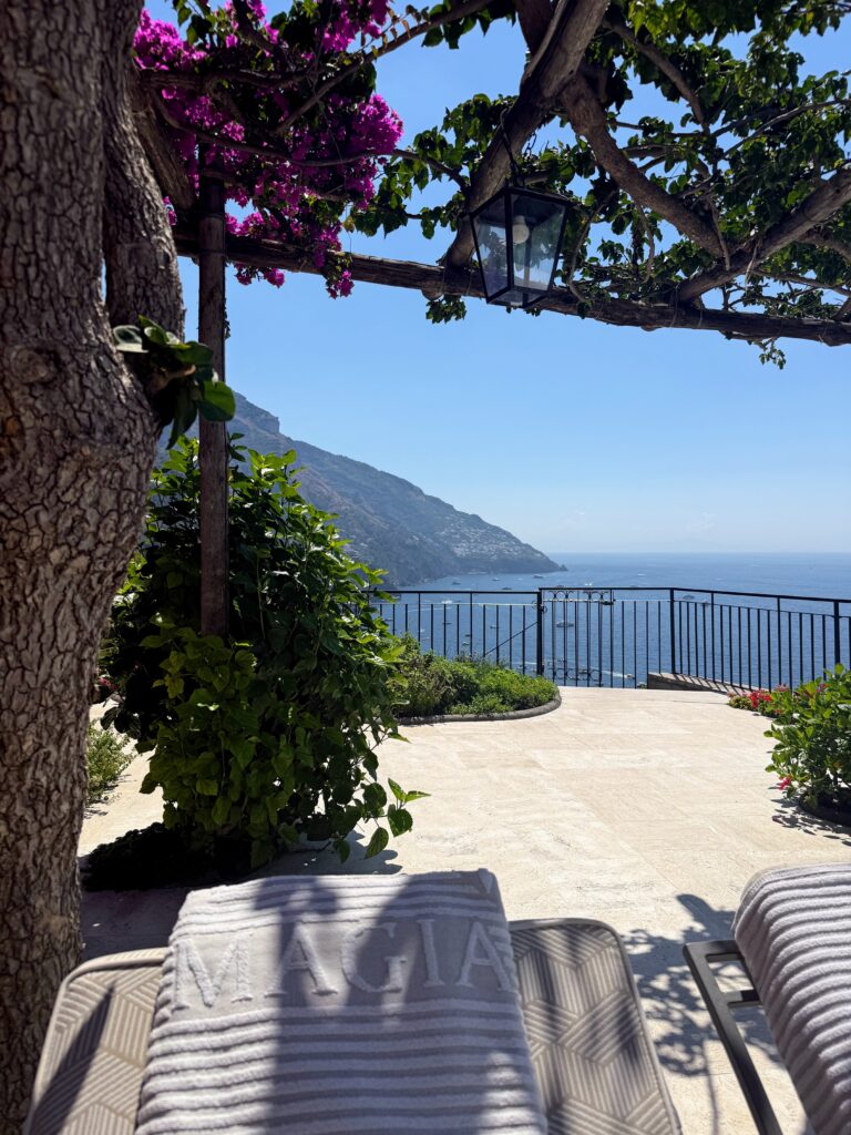 A large balcony overlooking the sea in Positano. A tree to the left with a trellis over head draped with purple flowers and a lantern.