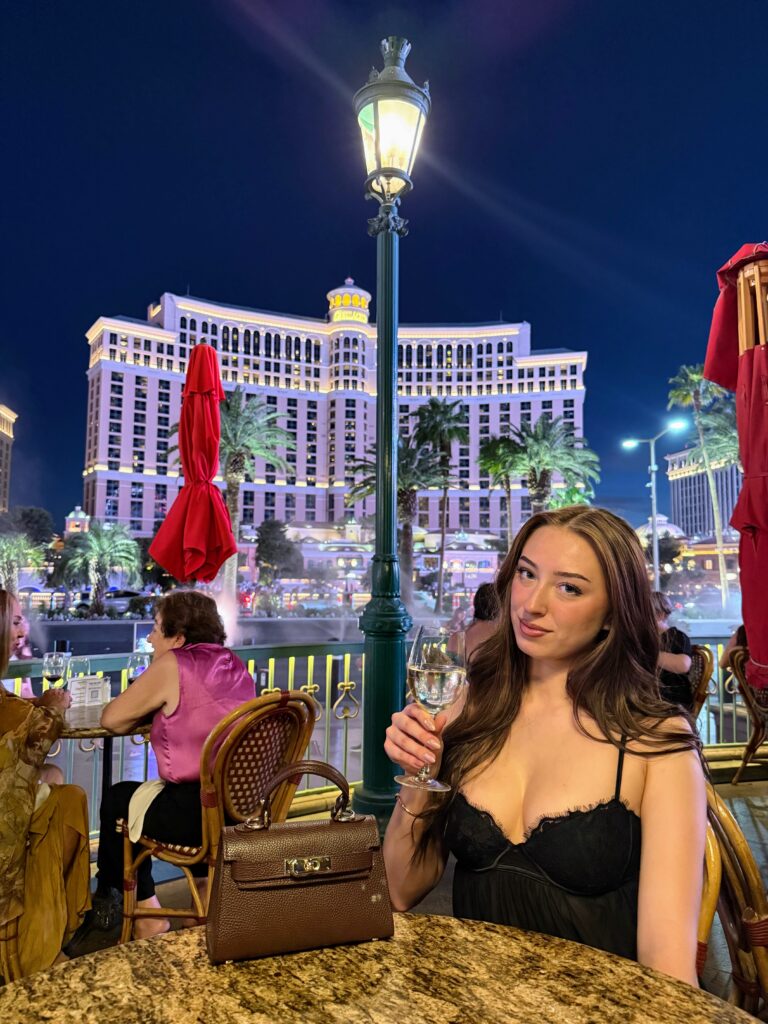 Brunette woman sitting with a glass of wine on a restaurant patio at night. Behind her across the street is the Bellagio hotel on the Las Vegas Strip.