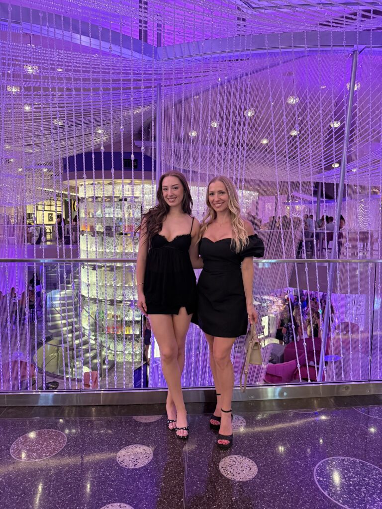 A blonde and brunette woman in front of the chandelier bar in Cosmopolitan hotel in Las Vegas. Purple lights with crystals hanging from floor to ceiling and a 3 story bar inside the giant chandelier of crystals.