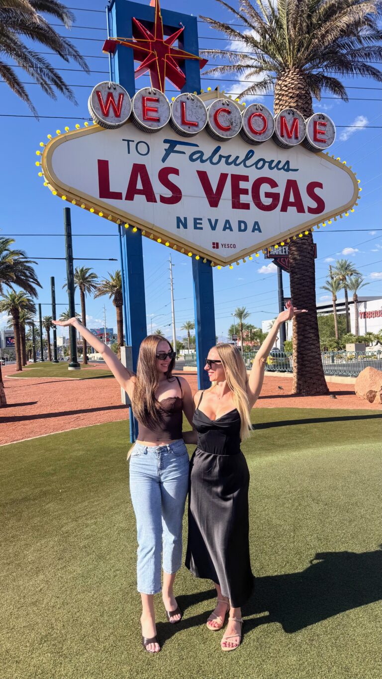 A blonde and brunette woman standing with their arms up in front of the Welcome to Fabulous Las Vegas sign.