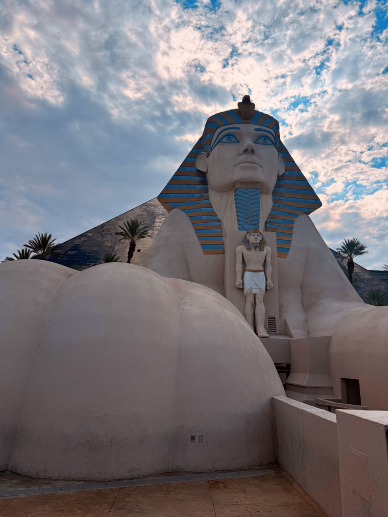 Exterior front of the Luxor hotel in Las Vegas. A large Sphinx with a black. pyramid and palm trees behind it.
