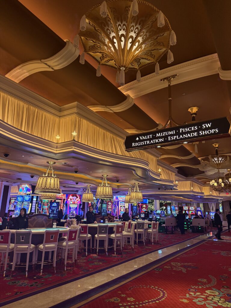 The casino floor at Wynn hotel. Red carpet with a floral pattern, chandeliers with lamp shades and tassels, and card game tables in front with slot machines in the distance.